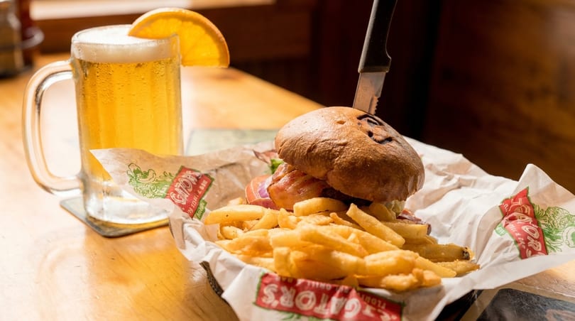 A burger with fries in a basket and a beer mug with an orange slice on a wooden table.