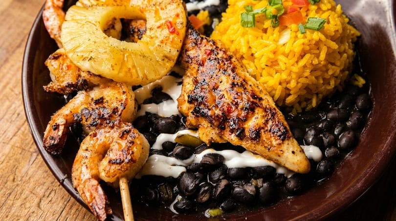 Grilled shrimp skewer, chicken, pineapple, yellow rice, and black beans with white sauce on a plate.