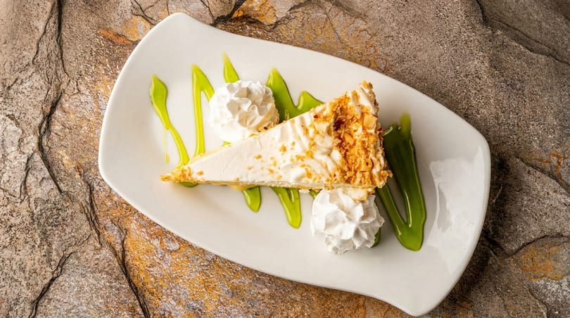 Slice of cheesecake with nuts, whipped cream, and green sauce on a white plate.