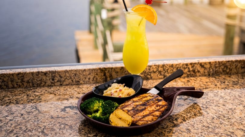Grilled fish with broccoli, mashed potatoes, and a cocktail on a granite counter overlooking water.